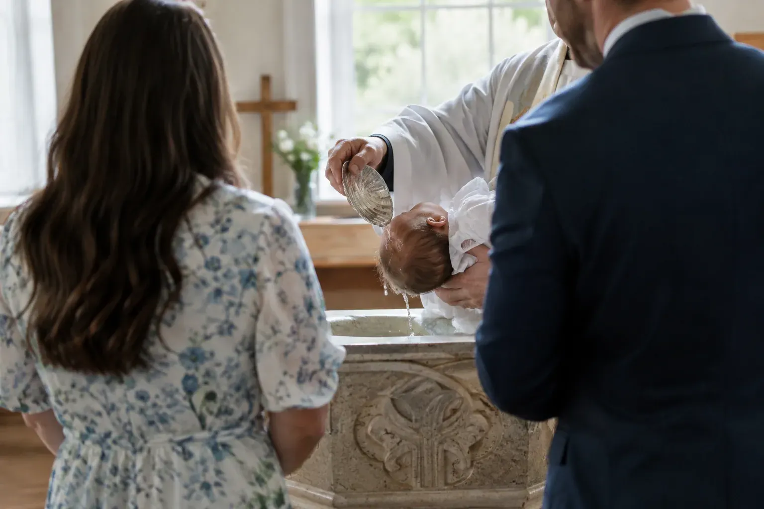 A minister pours water over an infant at a baptismal font as the family stands nearby.